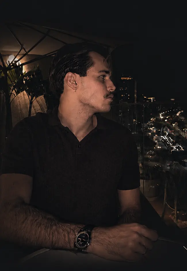 Tomás observando a cidade a partir de uma varanda à noite, com o trânsito iluminado ao fundo