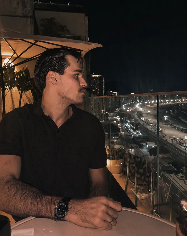 Tomás observando a cidade a partir de uma varanda à noite, com o trânsito iluminado ao fundo